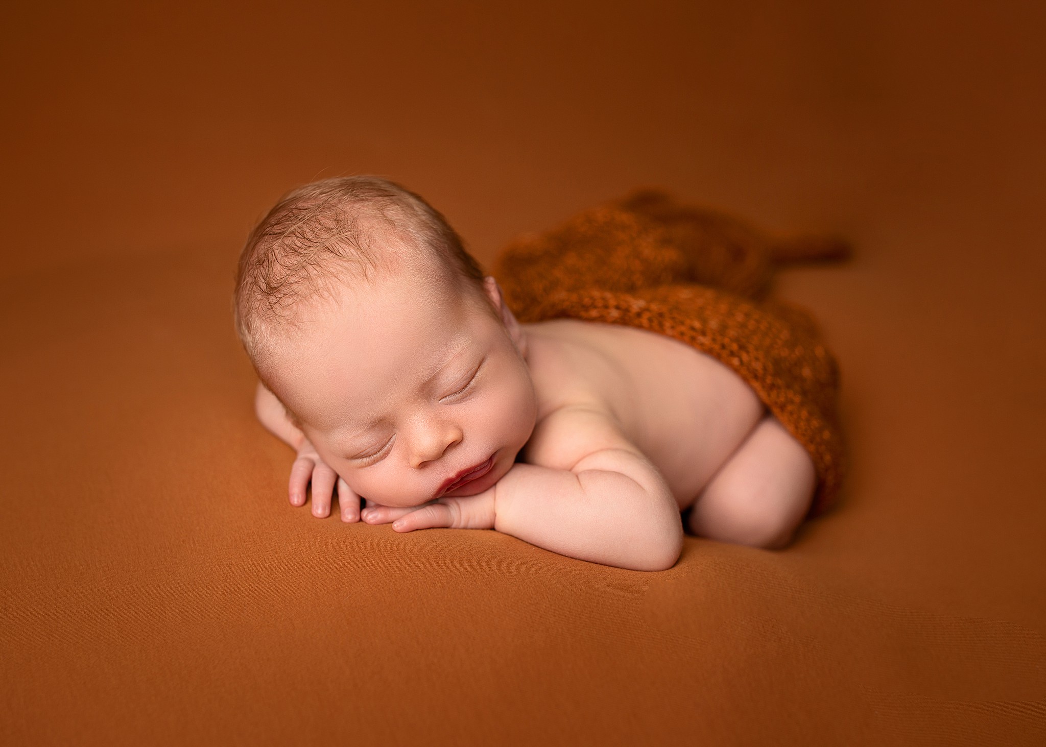 A Kamloops newborn photographer poses a sleeping baby boy with his head on his hands on a burnt orange fabric