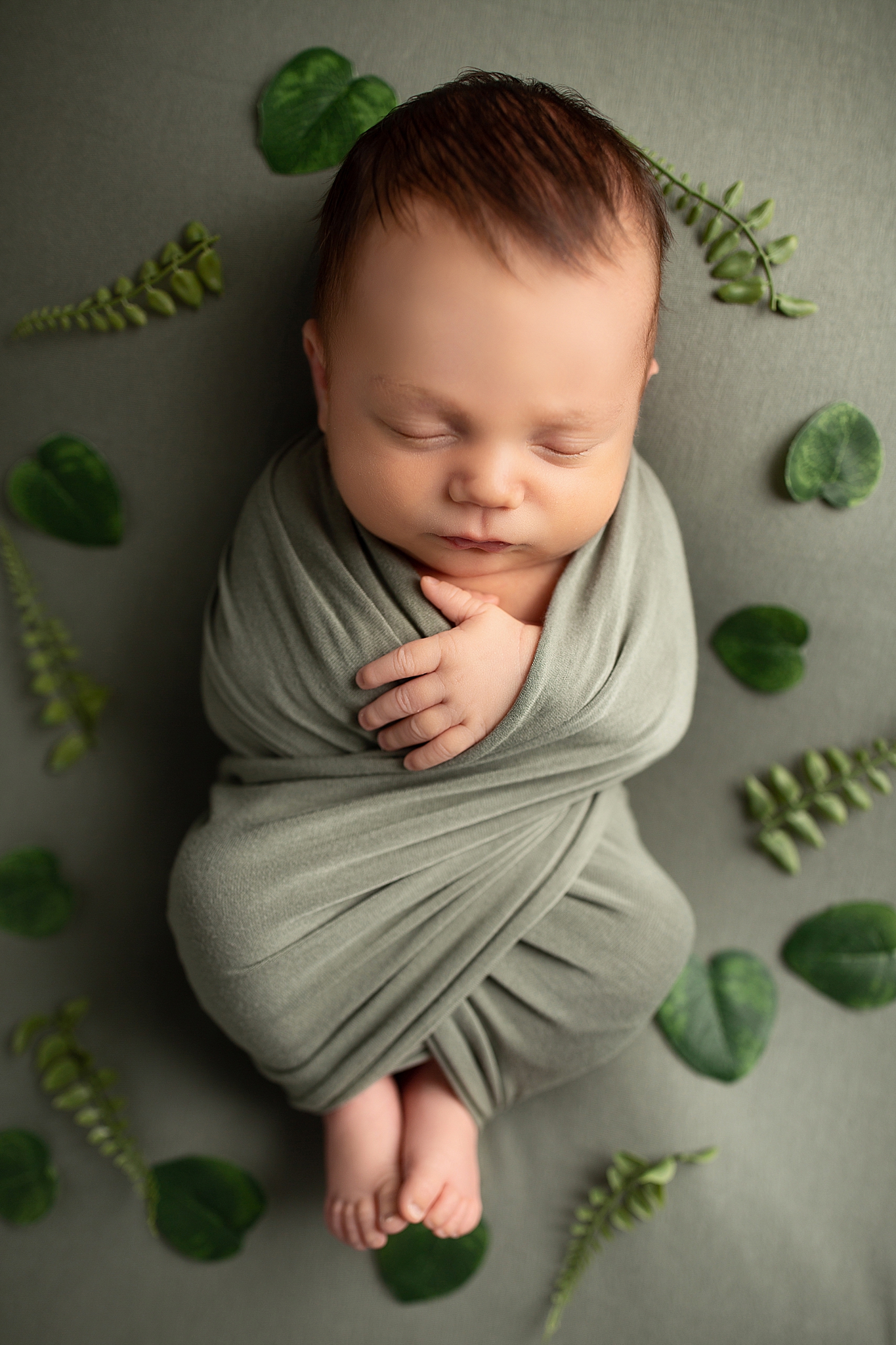 A newborn baby boy is wrapped up in a green wrap and photographed with green foliage around him.