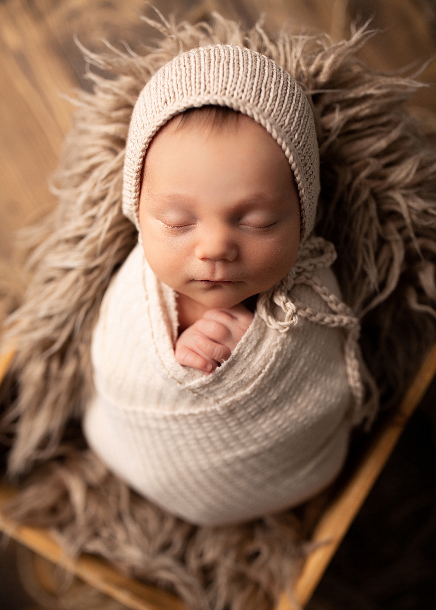 A newborn baby boy is photographed in a neutral beige coloured wrap and bonnet with wood flooring.